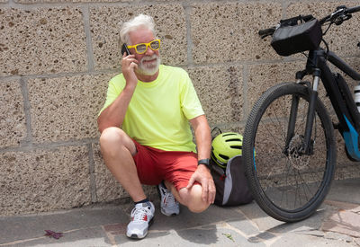 Bearded senior man answering mobile phone while crouching by bicycle against wall