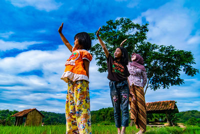 Low angle view of friends shielding eyes while standing on grassy field against sky