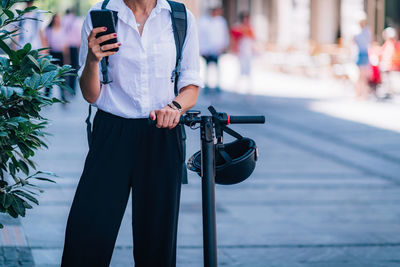 Midsection of woman using phone while standing with scooter on road