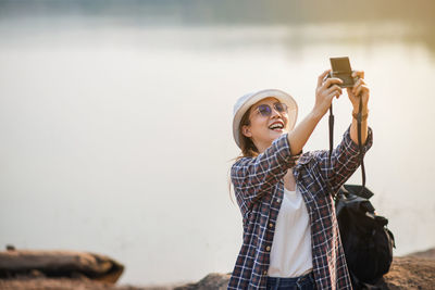 A female tourist happily takes a selfie by a reservoir with a camera.