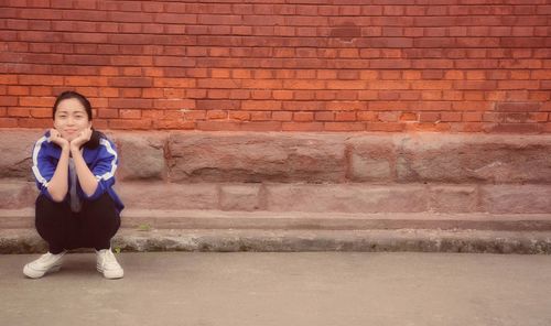 Portrait of young woman crouching on road against brick wall