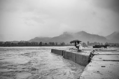 Man holding umbrella while crouching on pier over sea against sky during rainy season