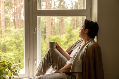 Middle age beautiful woman sitting on windowsill, drinking tea, dreaming. 50-year-old woman relaxing