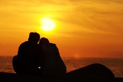 Rear view of couple sitting on coast against cloudy sky at dusk