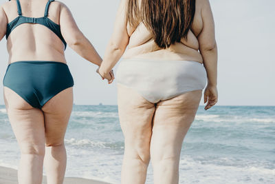 Back view of cropped unrecognizable curvy female friends in swimwear holding hands while walking in sea water during summer vacation