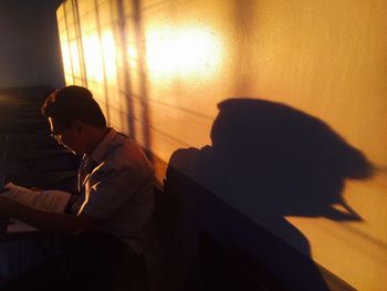 Side view of silhouette man sitting reading book while sitting by wall in room during sunset