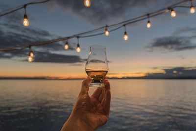 Close-up of human hand holding alcoholic drink by sea against sky during sunset