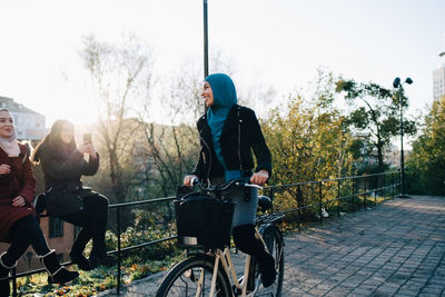 Smiling young woman cycling on footpath by female friends sitting on railing in city