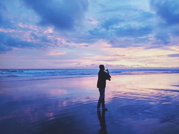 Woman photographing at beach against sky during sunset