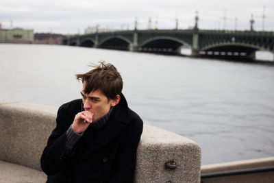 Man looking away while smoking cigarette against river and sky