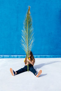 Teenager sitting with huge palm tree leaf on background of blue wall