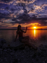 Man sitting on rock at beach against sky during sunset