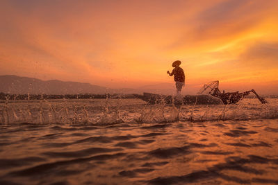 Fisherman throwing fishing net while standing on boat in sea against cloudy sky during sunset