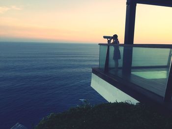 Woman looking at sea though telescope against sky during sunset