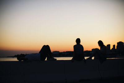 Silhouette friends on retaining wall against clear sky during sunset