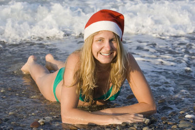 Portrait of happy young woman wearing santa hat lying on beach