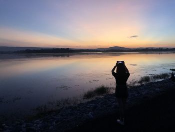 Rear view of silhouette woman photographing at lakeshore against sky during sunset