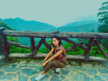 Portrait of young woman sitting on railing against mountain