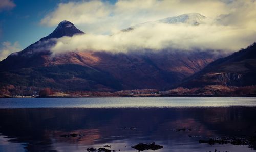 Scenic view of rocky mountains in front of lake against cloudy sky on sunny day