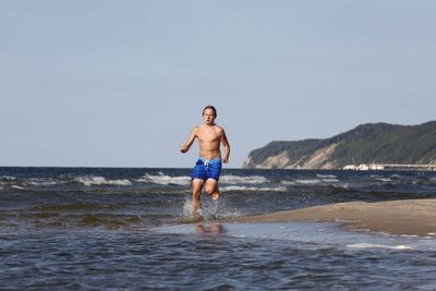 Shirtless young man running at sea shore against clear sky