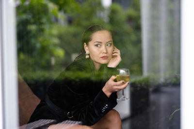 Portrait young woman sitting on armchair holding glass of wine in her hand shot through the glass