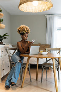 Full body of african american female freelancer sitting on chair and watching laptop while browsing on smartphone at table with breakfast