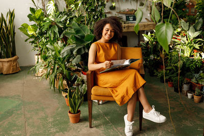 Smiling girl sitting with book on chair against plants