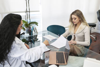 Young woman signing on paper sitting with doctor at desk in aesthetic clinic