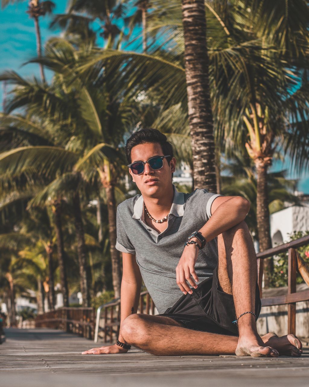 sunglasses, fashion, one person, young men, young adult, tropical climate, glasses, tree, front view, real people, palm tree, leisure activity, casual clothing, sitting, plant, lifestyles, day, portrait, nature, outdoors