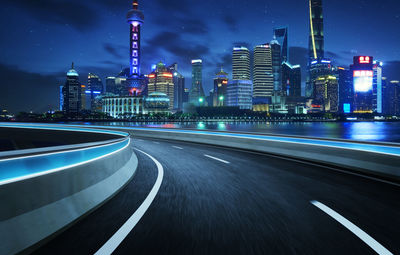 Light trails on road by illuminated buildings against sky at night