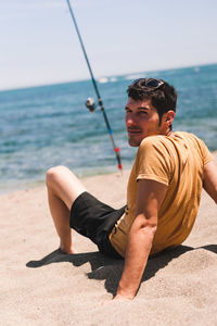 Young man sitting on shore at beach against sky