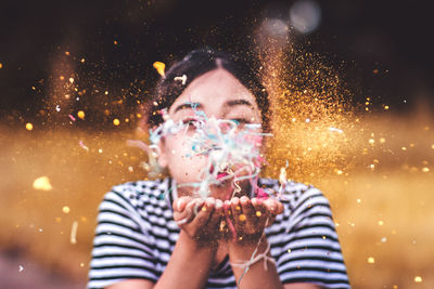 Close-up of smiling young woman blowing confetti while standing outdoors