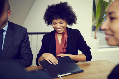 Smiling young female business lawyer holding file while sitting with colleagues at conference table in office during mee