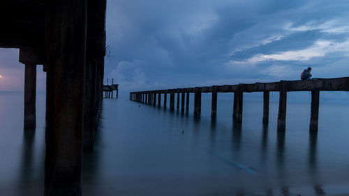 Man sitting on pier over sea against cloudy sky