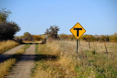 T intersection sign on grassy field by dirt road against blue sky