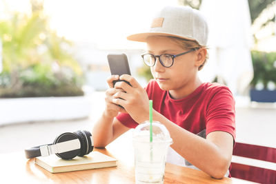 Boy wearing cap using smart phone with headphones and book on table while sitting at cafe