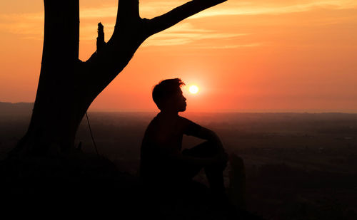 Silhouette man sitting against tree and sky during sunset