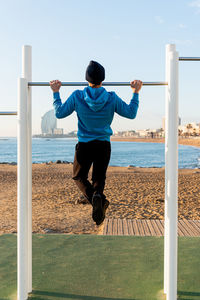 Full body back view of athletic male in warm activewear doing pull ups on metal bar while exercising on sports ground on urban beach