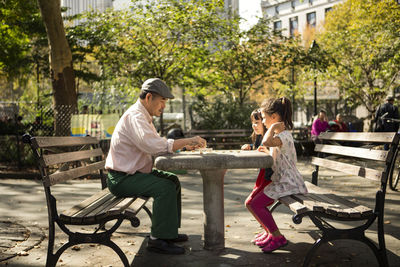 Side view of grandfather and granddaughters playing game while sitting on bench in park