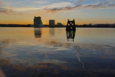 Silhouette people on sea by buildings against sky during sunset