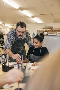 Instructor with paintbrush teaching student sitting at table in art class