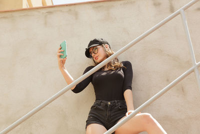 Young woman taking selfie standing against wall