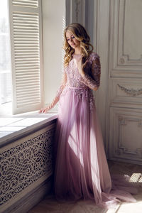 Beautiful young woman in pink lace dress standing next to the window in the white room