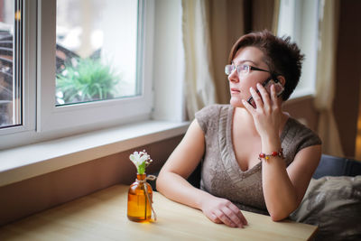 Woman answering smart phone while looking through window at home