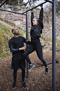 Mid adult man assisting woman exercising on monkey bars in forest