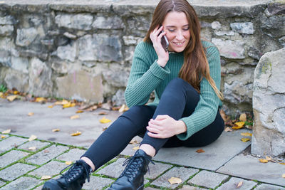 Portrait of a young caucasian woman with brown hair sitting with her back supported by a stone bridge and talking on her mobile phone. dressed in a green sweater, dark pants and military boot