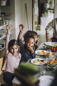 Happy girls and boys with hands raised at dining table during breakfast at kindergarten