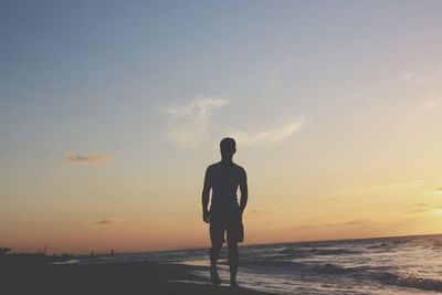 Rear view of silhouette man standing at beach against sky during sunset