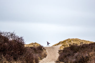 Woman jumping on dirt road amidst bush at hill against sky