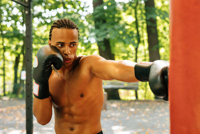 Rear view of shirtless man boxing while standing against tree